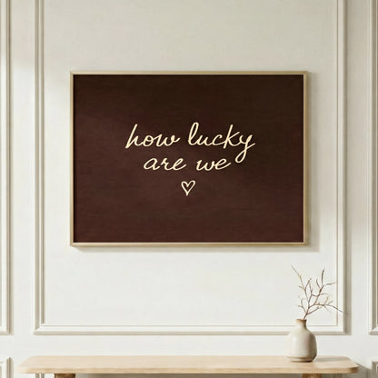 Horizontal "how lucky are we" typography poster in a light wood frame, dark brown aesthetic wall art over a minimalist console table