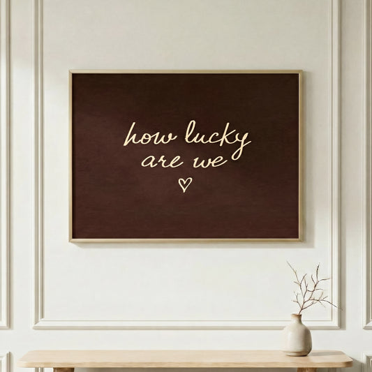 Horizontal "how lucky are we" typography poster in a light wood frame, dark brown aesthetic wall art over a minimalist console table