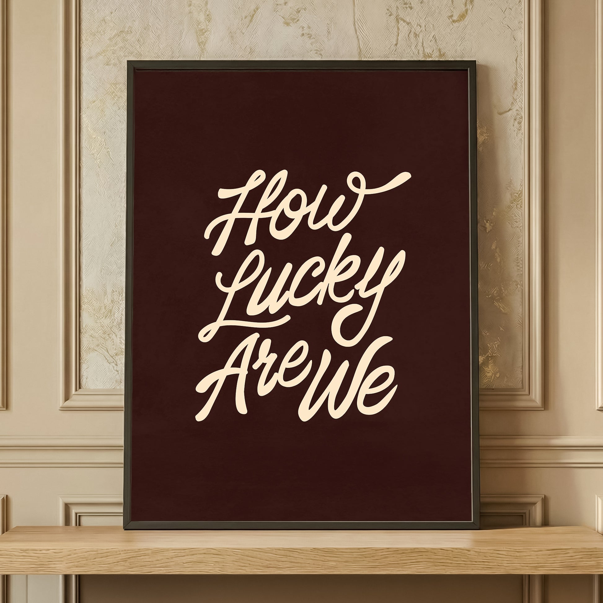 Framed 'How Lucky Are We' typography print in dark brown and cream, retro aesthetic wall art resting on a wooden shelf