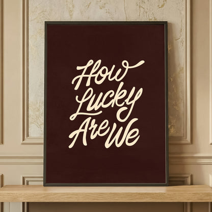 Framed 'How Lucky Are We' typography print in dark brown and cream, retro aesthetic wall art resting on a wooden shelf