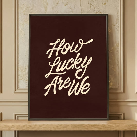 Framed 'How Lucky Are We' typography print in dark brown and cream, retro aesthetic wall art resting on a wooden shelf