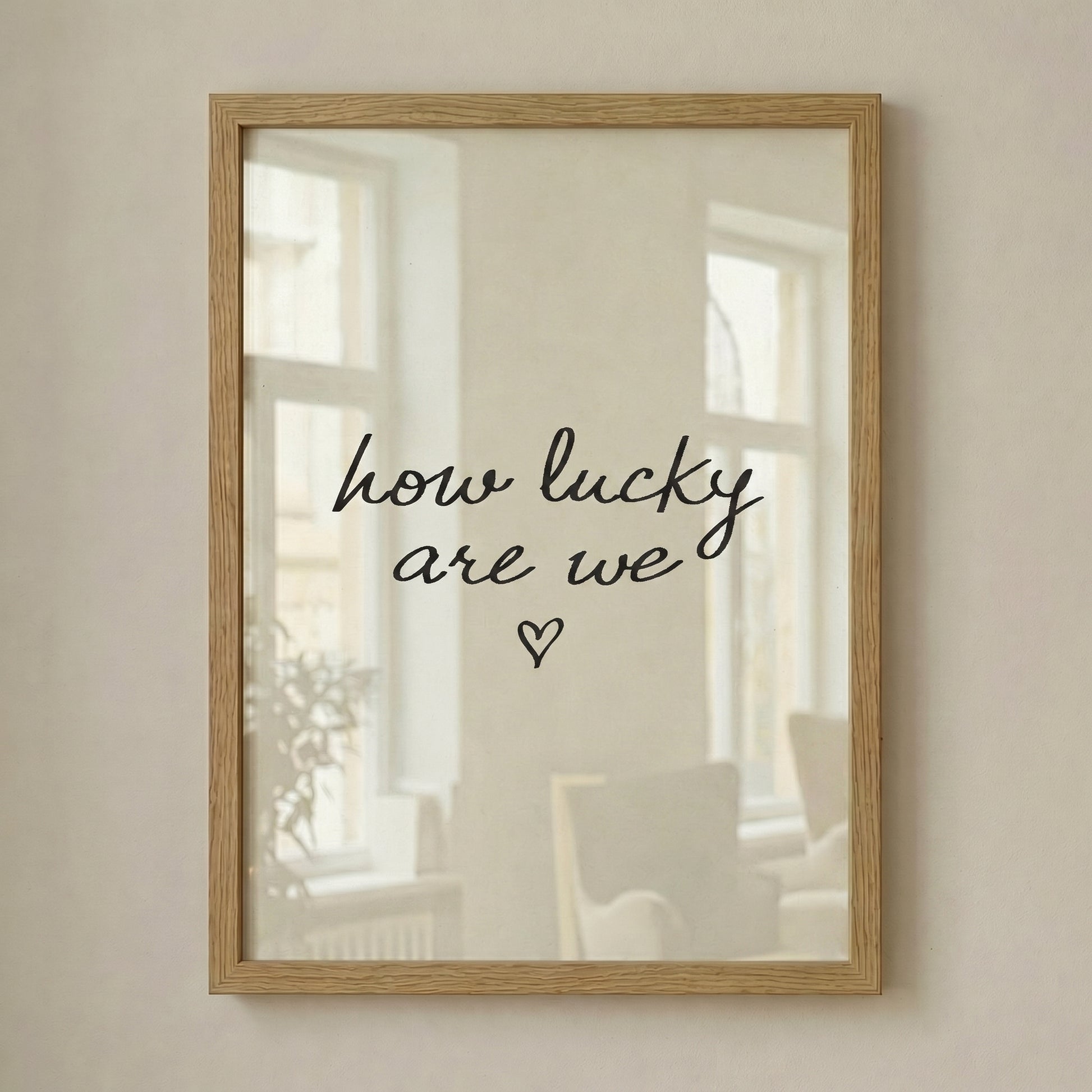Minimalist typography print saying how lucky are we with a small heart, placed in a light wood frame leaning against a white wall.