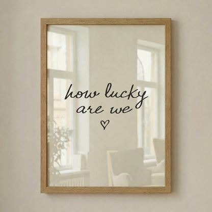 Minimalist typography print saying how lucky are we with a small heart, placed in a light wood frame leaning against a white wall.