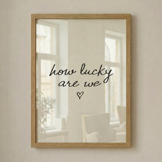 Minimalist typography print saying how lucky are we with a small heart, placed in a light wood frame leaning against a white wall.