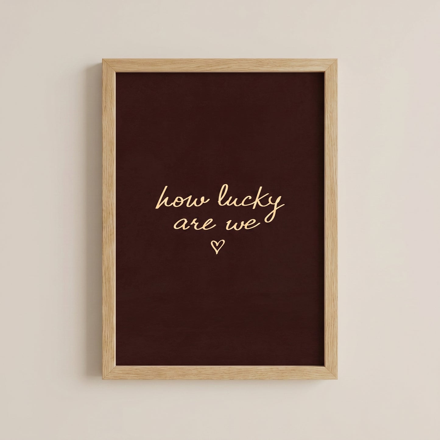 Minimalist typography print saying how lucky are we with a small heart on a dark brown background, placed in a light wood frame.