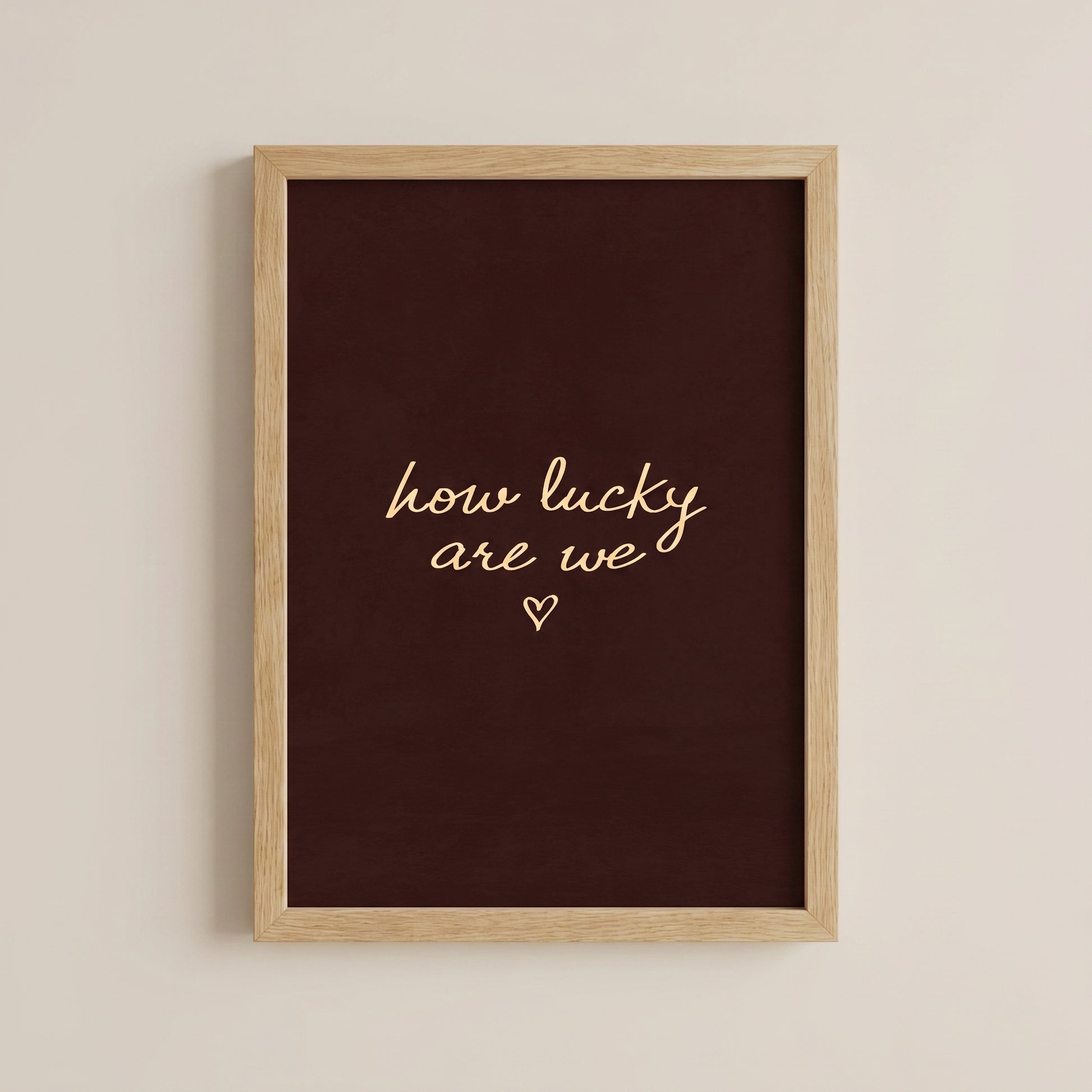 Minimalist typography print saying how lucky are we with a small heart on a dark brown background, placed in a light wood frame.
