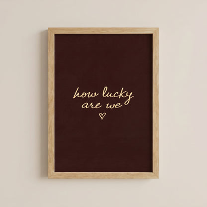 Minimalist typography print saying how lucky are we with a small heart on a dark brown background, placed in a light wood frame.