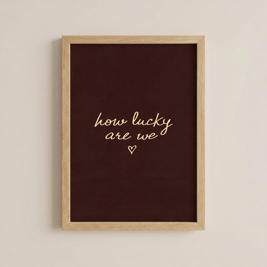 Minimalist typography print saying how lucky are we with a small heart on a dark brown background, placed in a light wood frame.