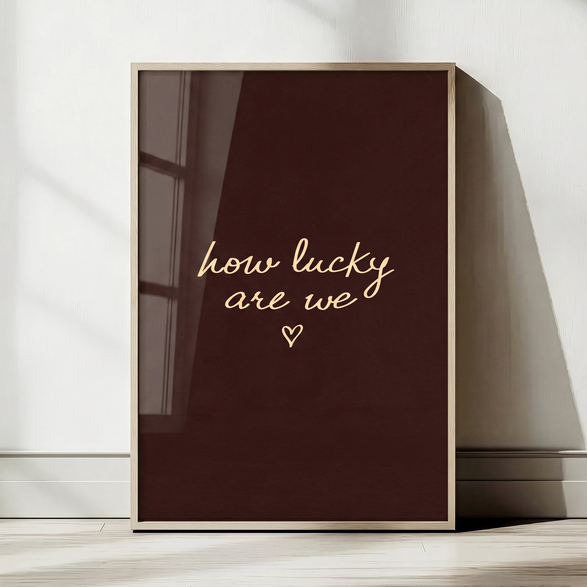 Minimalist typography print saying how lucky are we with a small heart on a dark brown background, placed in a light wood frame.