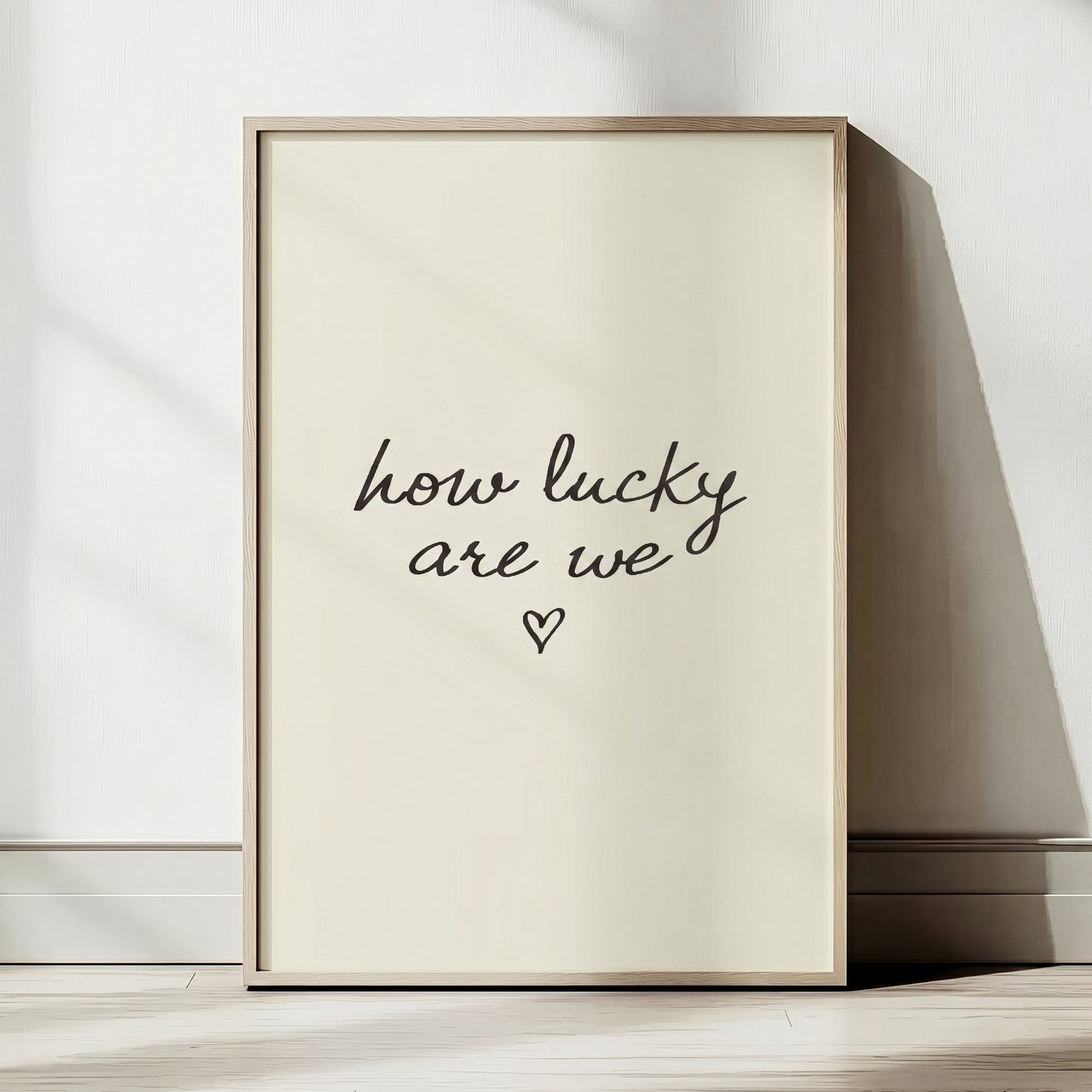 Minimalist typography print saying how lucky are we with a small heart, placed in a light wood frame leaning against a white wall.