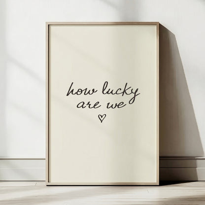 Minimalist typography print saying how lucky are we with a small heart, placed in a light wood frame leaning against a white wall.