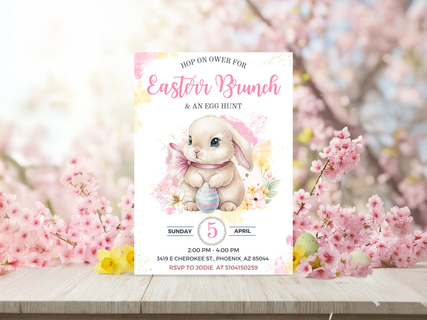 Watercolor Easter bunny holding a blue striped egg surrounded by pink cherry blossoms on a wooden table.