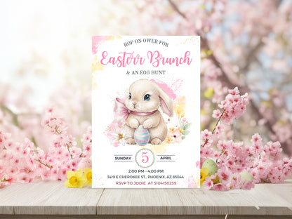 Watercolor Easter bunny holding a blue striped egg surrounded by pink cherry blossoms on a wooden table.