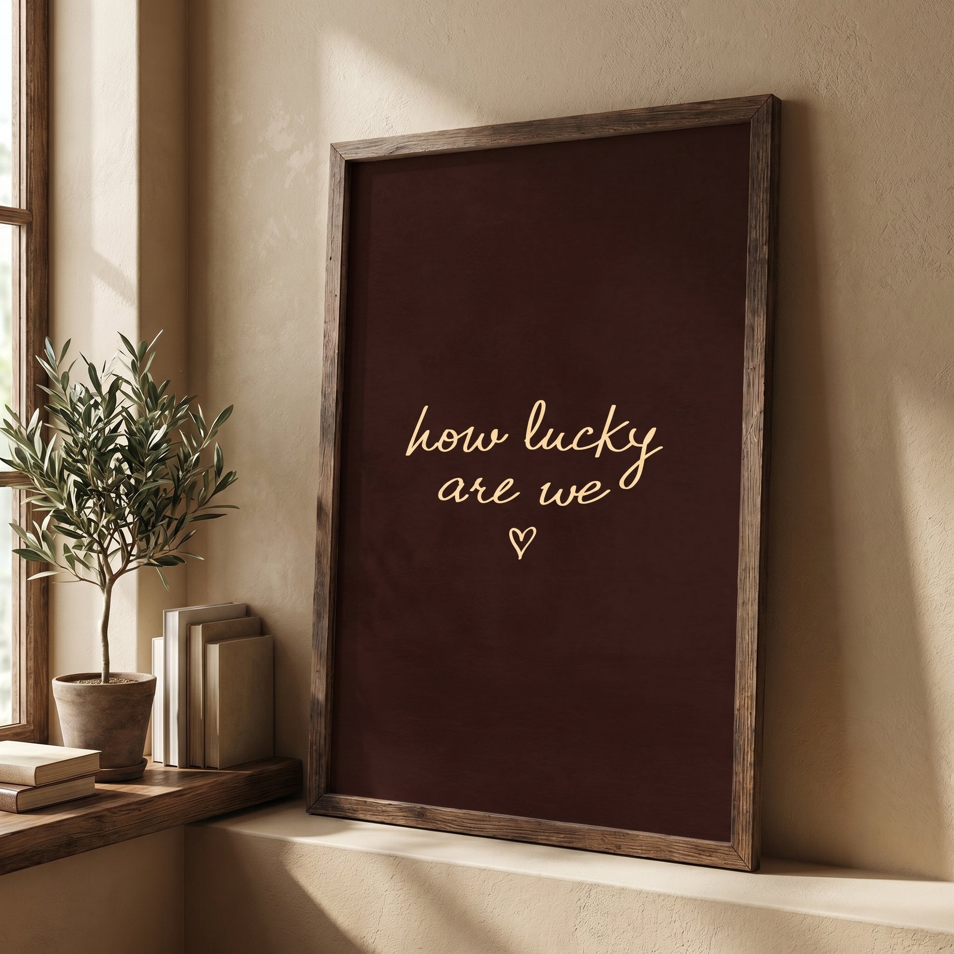 Framed moody dark brown typography poster leaning against a white wall next to a potted olive tree.