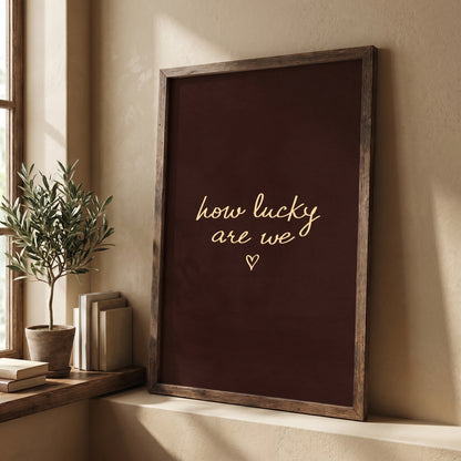 Framed moody dark brown typography poster leaning against a white wall next to a potted olive tree.
