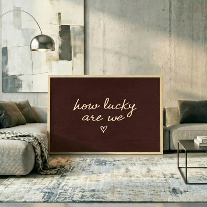 Large dark brown horizontal print reading "how lucky are we" leaning against a concrete wall in a modern living room