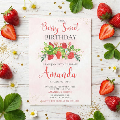 Printed strawberry theme first birthday invitation surrounded by fresh strawberries and green leaves.