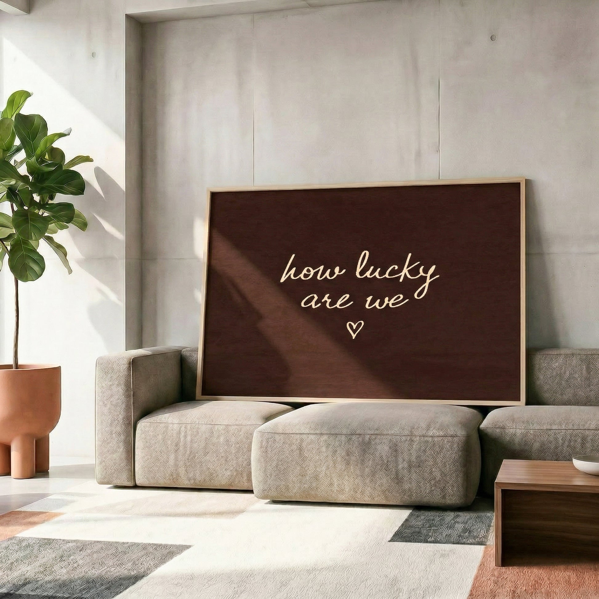Large dark brown horizontal print reading "how lucky are we" leaning against a concrete wall in a modern living room