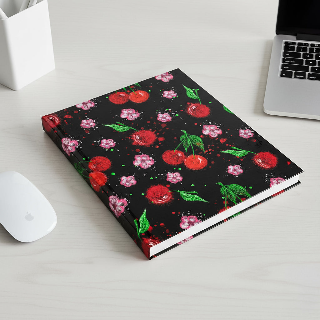 "A hardcover notebook with a cherry and flower pattern cover lying on a white desk next to a laptop."