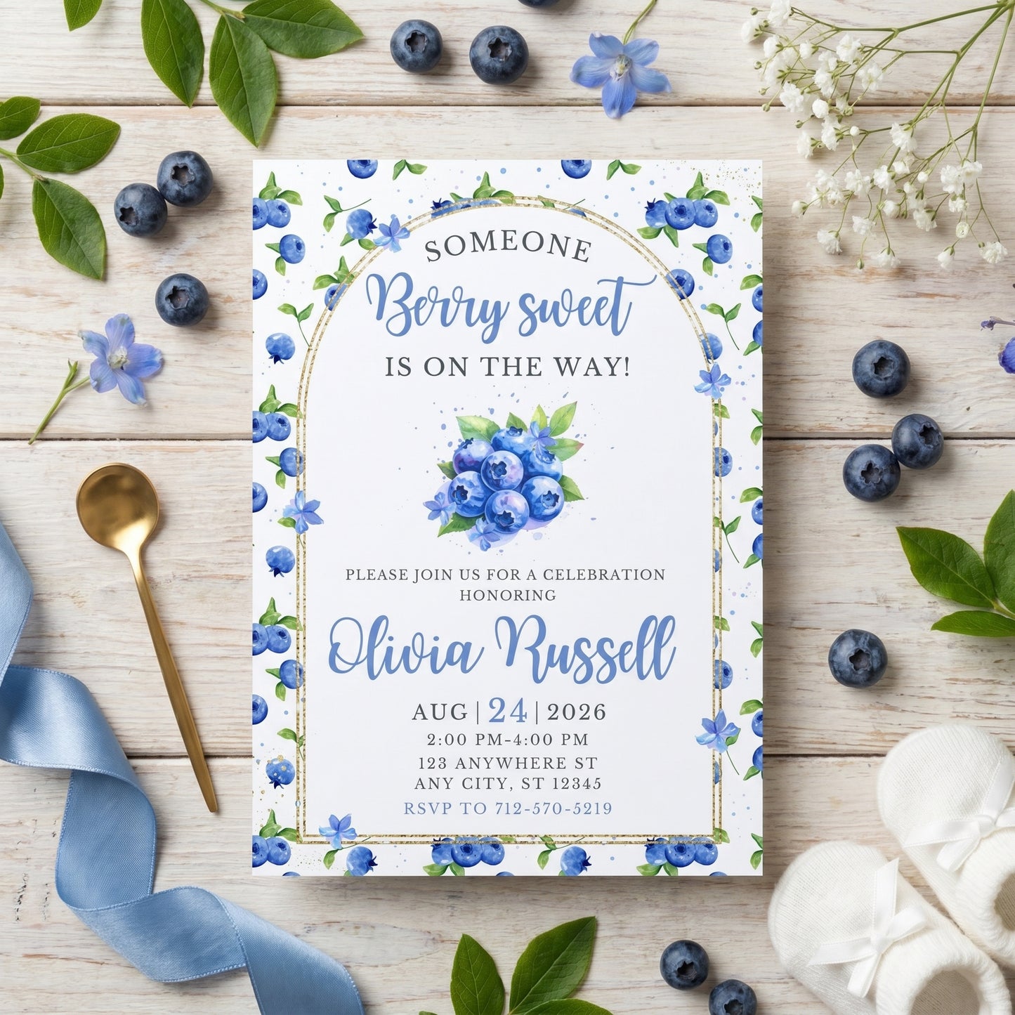 Printed blueberry baby shower invitation placed on a surface with fresh blueberries, blue ribbons, and white flowers