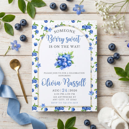 Printed blueberry baby shower invitation placed on a surface with fresh blueberries, blue ribbons, and white flowers