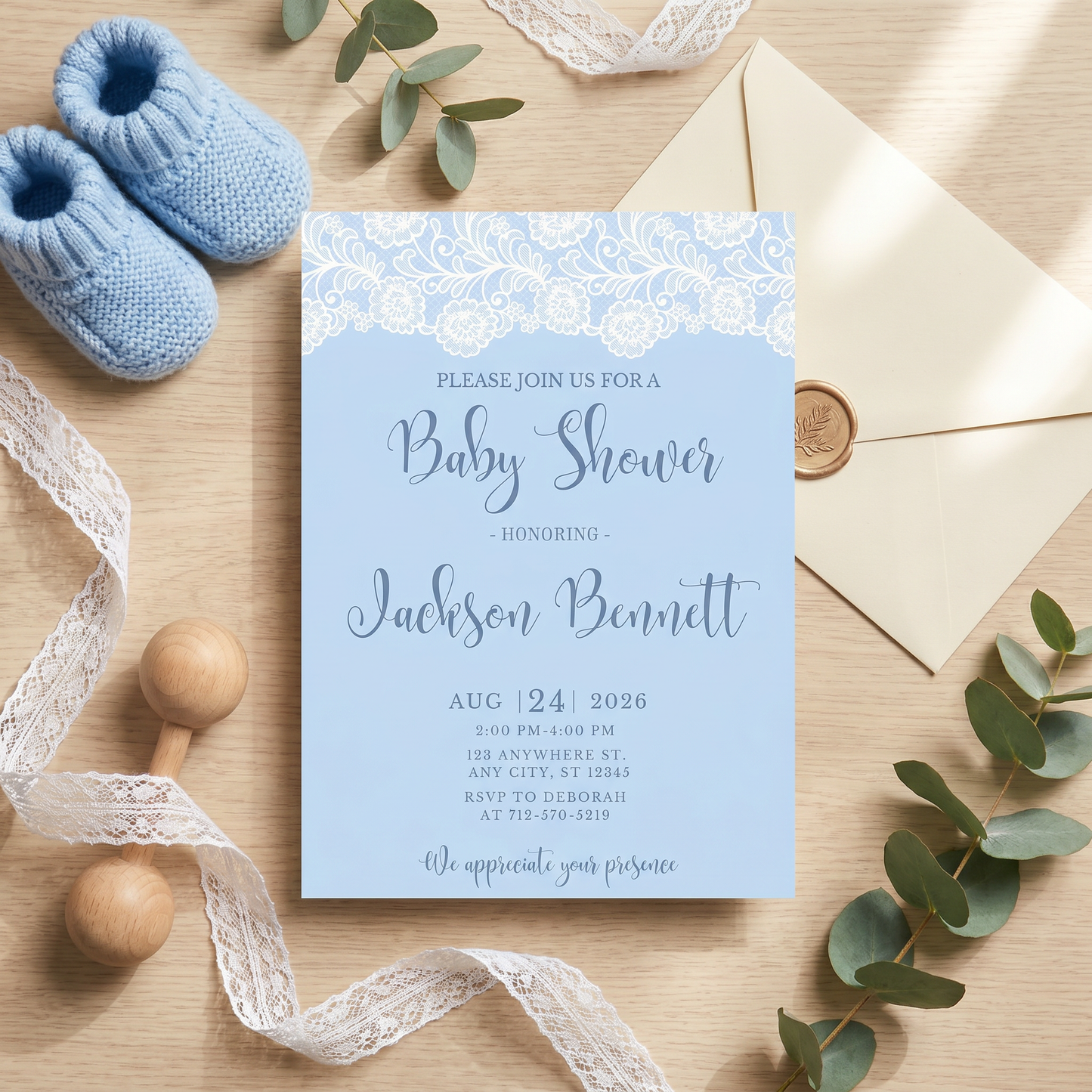 Printed blue lace baby shower invitation placed next to cute knitted baby boy booties and delicate dried flowers.