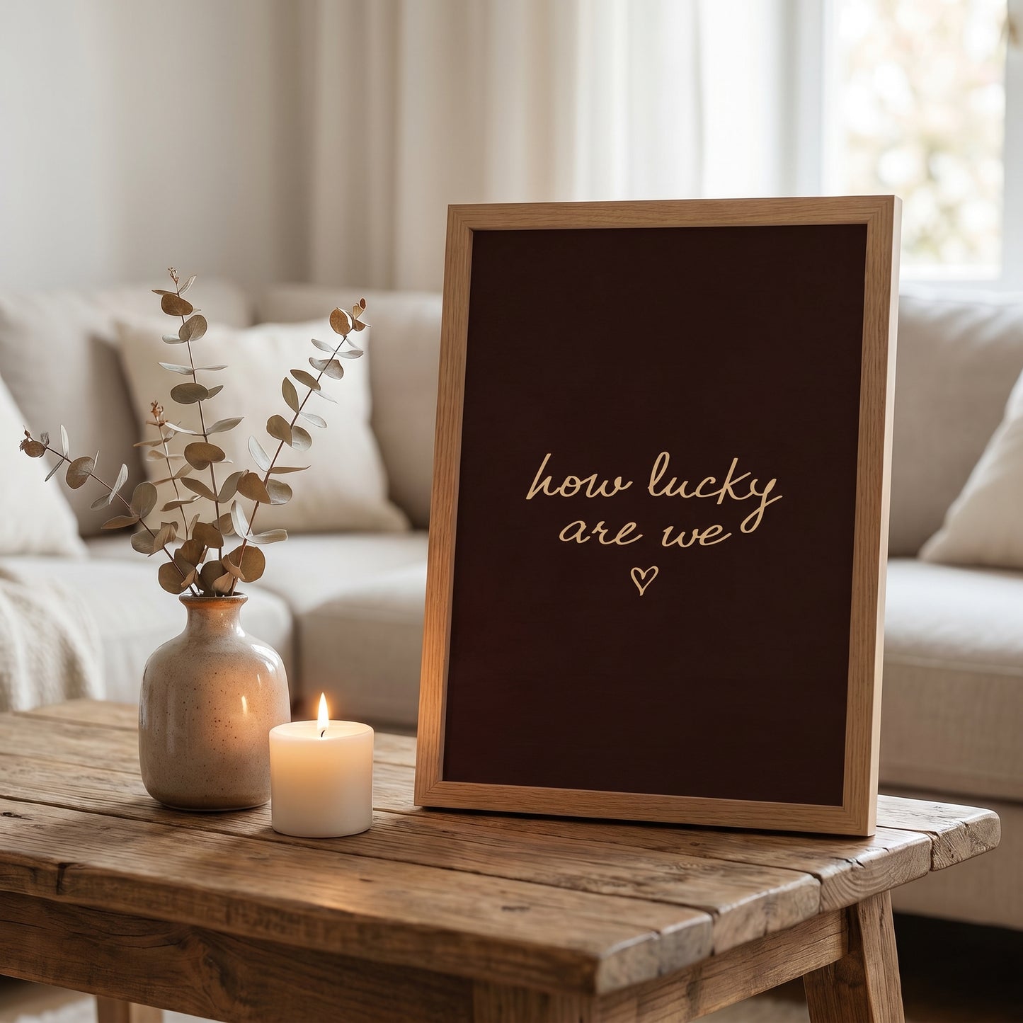 Cozy aesthetic room setup with a dark brown how lucky are we typography print, dried flowers, and a lit candle on a rustic wooden table.
